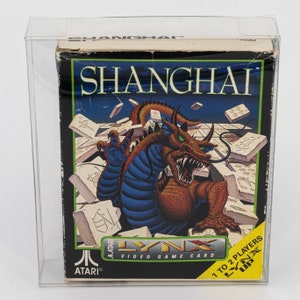 Può includere: Una scatola di gioco per Atari Lynx intitolata Shanghai, racchiusa in una custodia protettiva in plastica trasparente. La scatola presenta una grande illustrazione dettagliata di un drago tra tessere del mahjong, con il titolo Shanghai.
