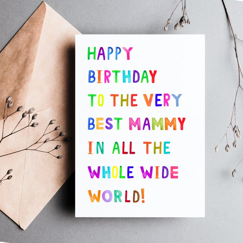 May include: A white birthday card with colorful text that reads "HAPPY BIRTHDAY TO THE VERY BEST MAMMY IN ALL THE WHOLE WIDE WORLD!" The card is next to a brown envelope and sprigs of greenery.