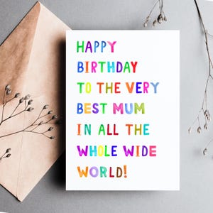 May include: A white birthday card with the text "HAPPY BIRTHDAY TO THE VERY BEST MUM IN ALL THE WHOLE WIDE WORLD!" in colorful, uppercase letters. The card is next to a brown envelope and sprigs of dried flowers.
