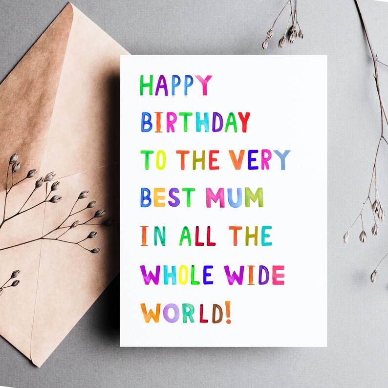 May include: A white birthday card with the text "HAPPY BIRTHDAY TO THE VERY BEST MUM IN ALL THE WHOLE WIDE WORLD!" in colorful, uppercase letters. The card is next to a brown envelope and sprigs of dried flowers.