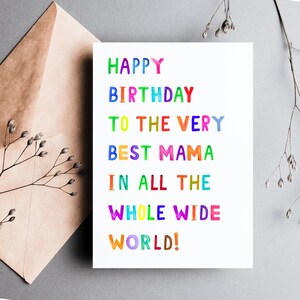 May include: A white birthday card with the text "HAPPY BIRTHDAY TO THE VERY BEST MAMA IN ALL THE WHOLE WIDE WORLD!" in colorful, watercolor-style lettering. The card is next to a brown envelope and sprigs of greenery.