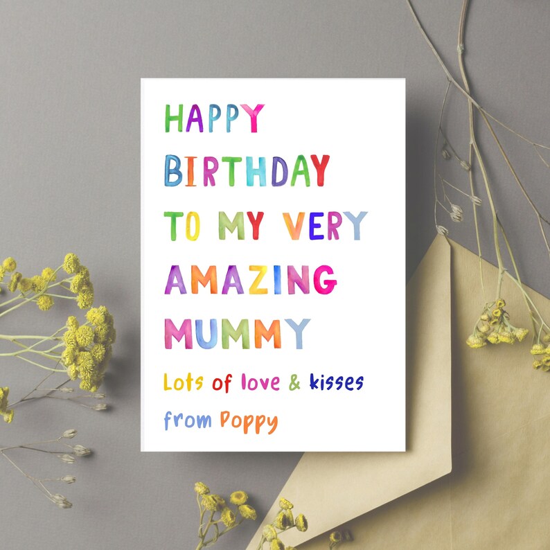 May include: A white greeting card with colorful text that reads "HAPPY BIRTHDAY TO MY VERY AMAZING MUMMY" in rainbow hues. The card also includes "Lots of love & kisses from Poppy" in blue and orange. A brown envelope and yellow flowers are in the background.