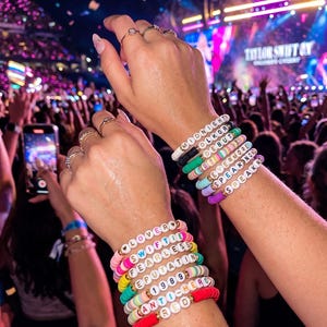 Könnte beinhalten: Zwei Handgelenke mit bunten Perlenarmbändern. Jedes Armband hat weiße Buchstabenperlen mit Wörtern wie "LOVER", "SWIFTIE" und "ANTI-HERO". Die Armbänder sind gestapelt und zeigen eine Vielzahl von Farben.