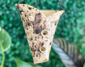 May include: A hanging planter made of ceramic with a speckled brown and white glaze. The planter has a cone shape with a wide opening at the top and a narrow opening at the bottom. There are several drainage holes in the sides of the planter.