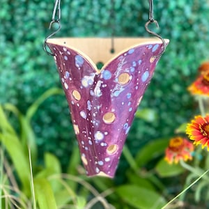 Puede incluir: Maceta colgante de cerámica morada con motas blancas y agujeros. La maceta tiene forma de cono y una abertura ancha en la parte superior. Está colgada de una cadena de metal negro.