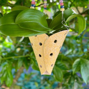 May include: A hanging yellow planter with a floral pattern and circular cutouts, suspended by beaded strands. The planter holds green leaves, set against a blurred green background. The beads are colorful, with shades of pink, yellow, and blue.