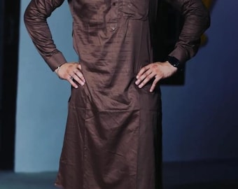 Pode incluir: Um homem vestindo uma kurta marrom de manga comprida com uma calça combinando. Ele está de pé com as mãos nos quadris. A kurta tem um colarinho abotoado e um bolso do lado esquerdo. O homem está usando sapatos dourados.