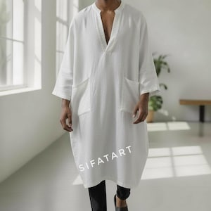Puede incluir: Una túnica de lino blanco con cuello en V y dos bolsillos delanteros. La túnica llega a la rodilla y se usa con pantalones negros y sandalias. El texto "SIFATART" está impreso en la túnica.