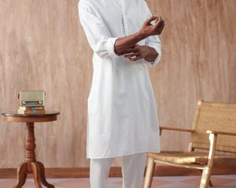 Pode incluir: Uma kurta branca de manga comprida com gola mandarim e fecho de botão. A kurta é combinada com calças brancas e sandálias marrons. O modelo está em pé em frente a uma parede castanho com uma cadeira e uma mesa de madeira.