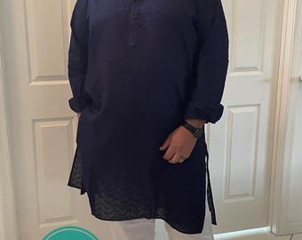 Può includere: Un uomo che indossa una kurta blu scuro a maniche lunghe con pantaloni bianchi. È in piedi davanti a una porta bianca con una cornice bianca. L'uomo sorride e ha la barba e gli occhiali.