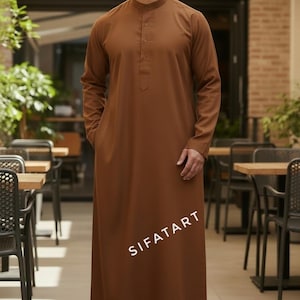 May include: A brown, long-sleeved thobe with a mandarin collar and buttoned placket. The garment, with the text "SIFATART" printed on the front, is worn by a person standing in a restaurant setting.