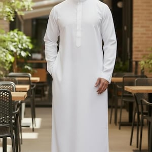May include: A white, long-sleeved thobe with a mandarin collar and a buttoned placket. The garment has a relaxed fit and pockets. The person is standing in an outdoor setting with tables and chairs.