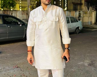 May include: A man wearing a white long-sleeved kurta and white pants with a red, orange, and yellow turban. He is standing in front of a building with lights strung across the front.
