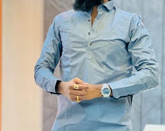 May include: A man wearing a light blue long-sleeved shirt with a button-up collar and a long hemline. He is wearing a silver watch on his left wrist and two gold rings on his right hand. The shirt has a small, dark blue design on the left side of the hemline. The man has a long, dark beard and is looking to the right.
