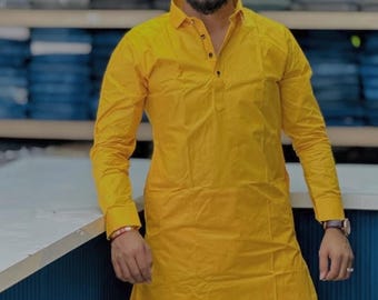 Puede incluir: Un hombre lleva una kurta amarilla de manga larga con cuello blanco abotonado y pantalones blancos. La kurta tiene un cierre de botón en el cuello. El hombre está de pie frente a una pared de jeans azules apilados.