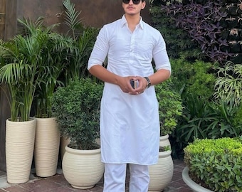 May include: A man wearing a white long-sleeved shirt and white pants with black loafers. He is standing in front of a green wall with plants and wearing sunglasses.