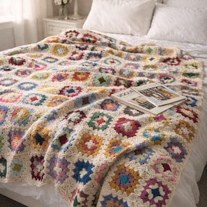 Op de afbeelding: Een handgemaakte, kleurrijke gehaakte deken met een bloemenpatroon, gedrapeerd over een bed. De deken heeft vierkanten in verschillende kleuren zoals blauw, rood, geel en groen. Een tijdschrift ligt erop.