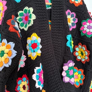 Puede incluir: Un c&aacute;rdigan negro de ganchillo adornado con coloridos cuadrados de la abuela florales. Las flores presentan una variedad de colores brillantes, incluyendo rosa, amarillo, azul y naranja. El c&aacute;rdigan tiene un cuello chal y un patr&oacute;n de puntada texturizada.