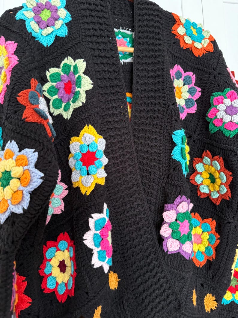 Puede incluir: Un c&aacute;rdigan negro de ganchillo adornado con coloridos cuadrados de la abuela florales. Las flores presentan una variedad de colores brillantes, incluyendo rosa, amarillo, azul y naranja. El c&aacute;rdigan tiene un cuello chal y un patr&oacute;n de puntada texturizada.