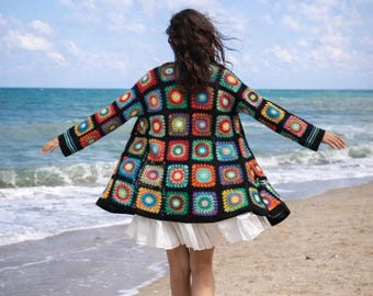 May include: A long-sleeved cardigan made of black yarn featuring a vibrant granny square pattern with multicolored centers in shades of red, orange, yellow, green, blue, and purple. The garment is worn over a white skirt with a ruffled hemline.