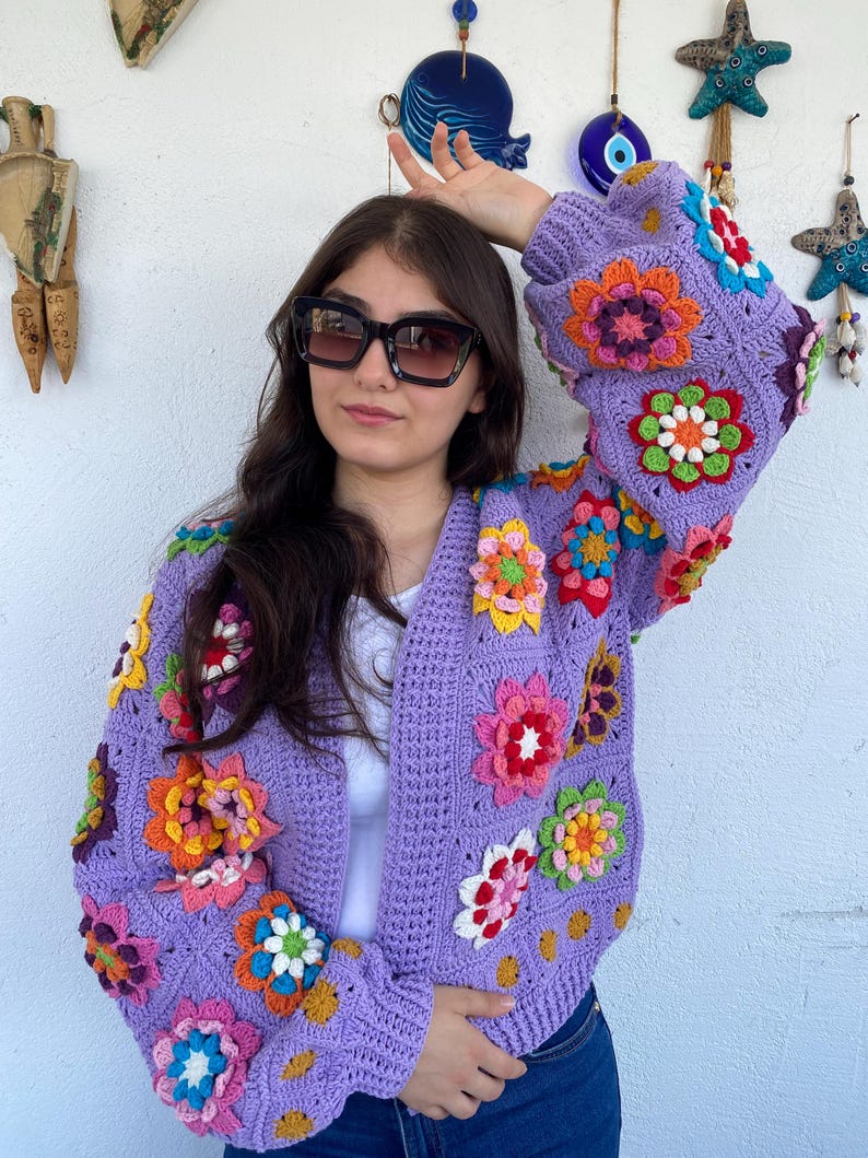 Puede incluir: Un c&aacute;rdigan de crochet lila con aplicaciones florales coloridas. El su&eacute;ter de frente abierto presenta paneles cuadrados adornados con flores vibrantes hechas a mano en tonos de rosa, amarillo, naranja y azul. El c&aacute;rdigan es una prenda &uacute;nica, hecha a mano.