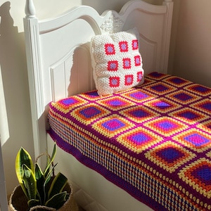 May include: A white bed with a crocheted throw blanket in shades of purple, orange, and yellow. The blanket has a square pattern. A white crocheted pillow with pink and purple squares is on the bed.