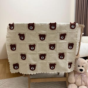 Può includere: Una coperta all'uncinetto color crema caratterizzata da un motivo a griglia ripetuto di blocchi quadrati. Ogni blocco contiene una piccola applicazione a forma di testa d'orso marrone scuro con dettagli bianchi su muso e orecchie, con bordo smerlato.