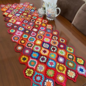 以下が含まれることがあります： 幾何学模様のカラフルな編み物のテーブルランナー。ランナーは赤、ピンク、青、緑、黄、オレンジの糸でできています。木製のテーブルの上に置かれています。