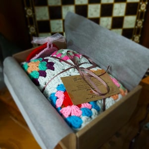 Pu&ograve; includere: Una colorata coperta all'uncinetto con motivi floreali in una scatola di cartone, legata con un nastro marrone e un piccolo cartellino. La coperta presenta un mix di colori tra cui blu, rosa, arancione e verde. La scatola &egrave; foderata con carta bianca.
