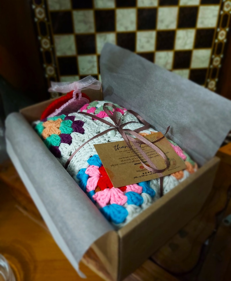 Pu&ograve; includere: Una colorata coperta all'uncinetto con motivi floreali in una scatola di cartone, legata con un nastro marrone e un piccolo cartellino. La coperta presenta un mix di colori tra cui blu, rosa, arancione e verde. La scatola &egrave; foderata con carta bianca.