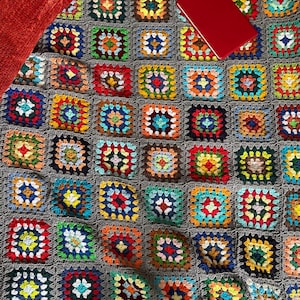 May include: A colourful crocheted blanket with a red notebook on top. The blanket is made of squares of different colours, including red, orange, yellow, green, blue, purple, and pink.