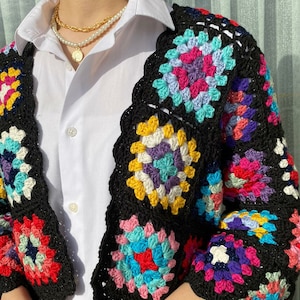 Könnte beinhalten: Eine schwarze Häkeljacke mit bunten Granny Squares. Die Jacke hat eine verkürzte Länge und Dreiviertelärmel.