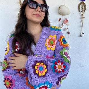 Puede incluir: Un c&aacute;rdigan de ganchillo color lavanda con un patr&oacute;n vibrante de cuadrados de la abuela florales multicolores. La prenda tiene mangas largas y cuello en V, confeccionada con hilo morado con flores en tonos amarillo, rosa, azul, naranja y verde.