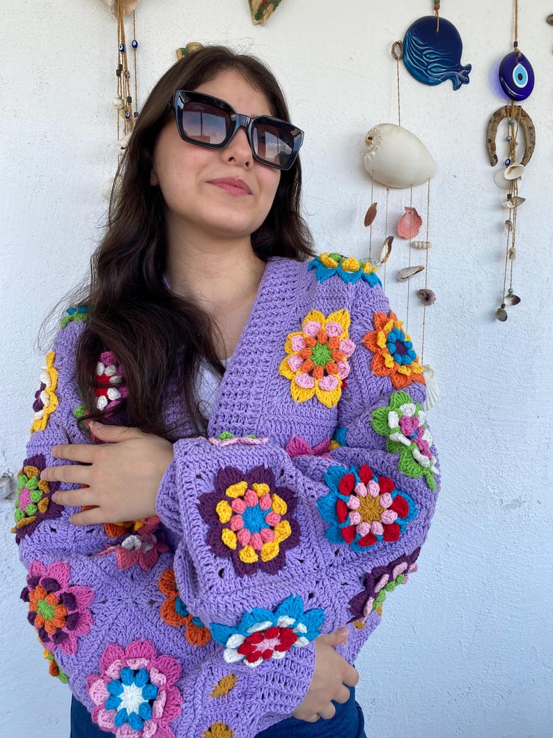 Puede incluir: Un c&aacute;rdigan de ganchillo color lavanda con un patr&oacute;n vibrante de cuadrados de la abuela florales multicolores. La prenda tiene mangas largas y cuello en V, confeccionada con hilo morado con flores en tonos amarillo, rosa, azul, naranja y verde.
