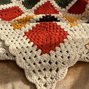 Pu&ograve; includere: Una coperta all'uncinetto fatta a mano con un design patchwork. La coperta presenta quadrati color crema, arancione, verde, bordeaux e giallo. Il lavoro all'uncinetto &egrave; dettagliato, con un bordo smerlato.