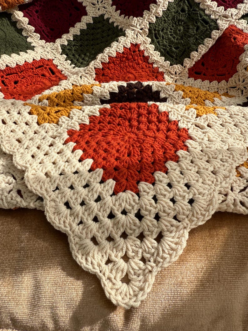 Pu&ograve; includere: Una coperta all'uncinetto fatta a mano con un design patchwork. La coperta presenta quadrati color crema, arancione, verde, bordeaux e giallo. Il lavoro all'uncinetto &egrave; dettagliato, con un bordo smerlato.