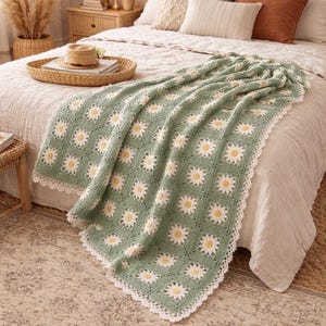 Op de afbeelding: Een gehaakte deken met een madeliefpatroon, met een lichtgroene achtergrond en witte en gele bloemdetails. De deken heeft een witte, geschulpte rand en is over een bed gedrapeerd. De algehele esthetiek is gezellig en handgemaakt.