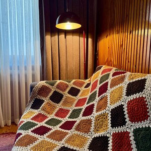 Pu&ograve; includere: Una coperta all'uncinetto fatta a mano con un motivo a rombi geometrici nei toni del marrone, verde, rosso e giallo. La coperta &egrave; drappeggiata su un divano grigio. I bordi sono rifiniti con un bordo bianco smerlato.