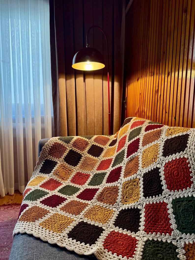Pu&ograve; includere: Una coperta all'uncinetto fatta a mano con un motivo a rombi geometrici nei toni del marrone, verde, rosso e giallo. La coperta &egrave; drappeggiata su un divano grigio. I bordi sono rifiniti con un bordo bianco smerlato.