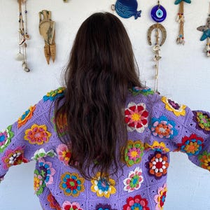 Puede incluir: Un su&eacute;ter de crochet morado con dise&ntilde;os de flores de colores. El su&eacute;ter tiene mangas largas y un ajuste holgado.