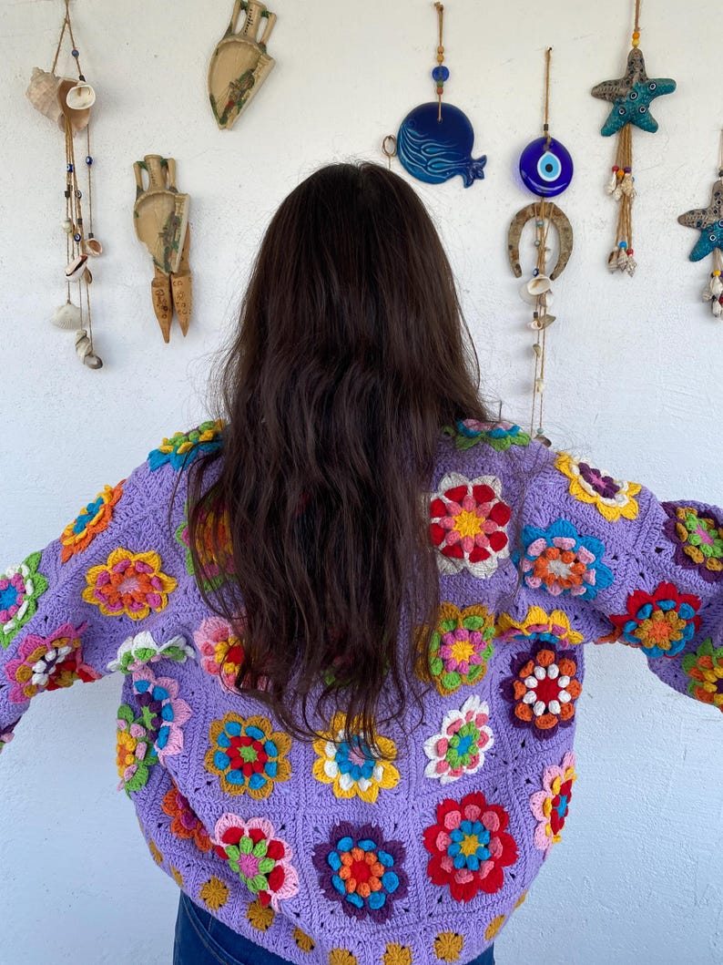 Puede incluir: Un su&eacute;ter de crochet morado con dise&ntilde;os de flores de colores. El su&eacute;ter tiene mangas largas y un ajuste holgado.
