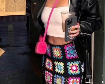 May include: A woman wearing a black leather jacket, a white crop top, and a colourful crochet mini skirt. She is holding a cup of coffee and a black phone case with the letters "NY" on it.