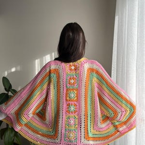 May include: A colorful, handmade crochet shawl in shades of pink, orange, green, and yellow. The shawl features a square design with floral accents and a striped pattern. The garment is worn over a white shirt and black shorts.