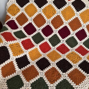 Pu&ograve; includere: Una coperta all'uncinetto fatta a mano con un design patchwork di quadrati in vari colori caldi, tra cui arancione, giallo, marrone e verde. I quadrati sono bordati da un bordo bianco e merlettato, creando un aspetto accogliente e strutturato. La coperta &egrave; drappeggiata su una sedia.