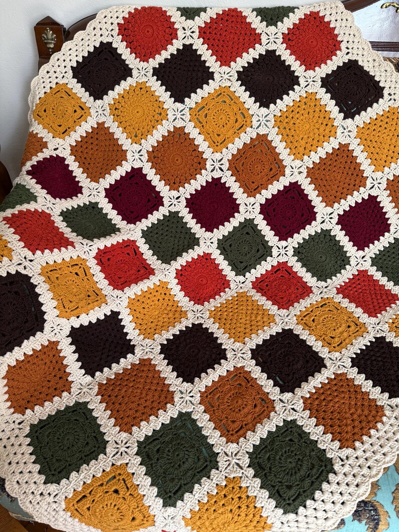 Pu&ograve; includere: Una coperta all'uncinetto fatta a mano con un design patchwork di quadrati in vari colori caldi, tra cui arancione, giallo, marrone e verde. I quadrati sono bordati da un bordo bianco e merlettato, creando un aspetto accogliente e strutturato. La coperta &egrave; drappeggiata su una sedia.