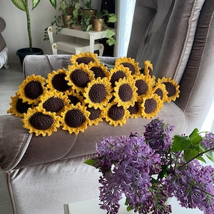 May include: A bouquet of crocheted sunflowers in shades of brown and yellow, arranged in a pile on a light brown armchair. A vase of purple lilac flowers sits on a white table in the foreground.
