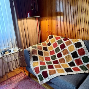Pu&ograve; includere: Una coperta all'uncinetto fatta a mano con un design patchwork in vari colori, tra cui rosso, verde, giallo e marrone. La coperta &egrave; drappeggiata su un divano grigio. Il bordo della coperta &egrave; bianco.