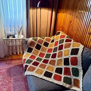 Pu&ograve; includere: Una coperta all'uncinetto fatta a mano con un motivo geometrico nei toni del rosso, arancione, giallo, verde e marrone. La coperta &egrave; drappeggiata su un divano grigio. I bordi sono rifiniti con un bordo color crema.