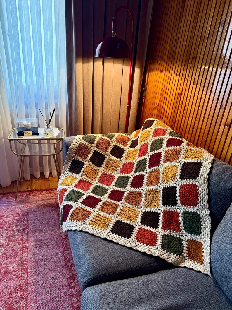 Pu&ograve; includere: Una coperta all'uncinetto fatta a mano con un motivo geometrico nei toni del rosso, arancione, giallo, verde e marrone. La coperta &egrave; drappeggiata su un divano grigio. I bordi sono rifiniti con un bordo color crema.