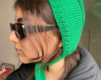 May include: A green knitted hat with a long tie. The hat is pulled down over the person's head and the tie is wrapped around their neck.